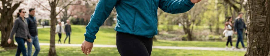 A smiling woman power walks along a park path beneath blossoming cherry blossom trees.