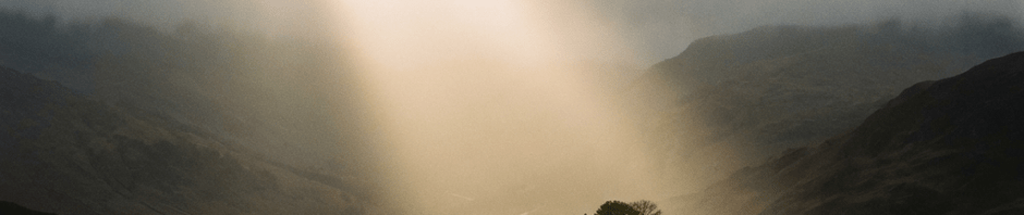 Sunbeam breaking through dark clouds to illuminate a stone circle and tree on a hill.
