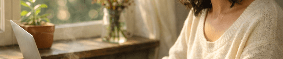 Woman typing on laptop at wooden table near window with tea cup and notebook