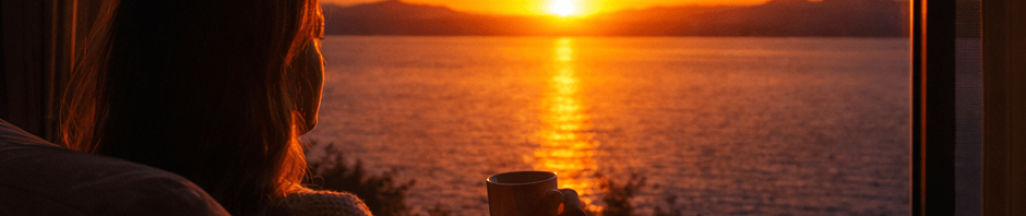 Person sitting on a chair by a window holding a cup, watching a vibrant sunset over a body of water