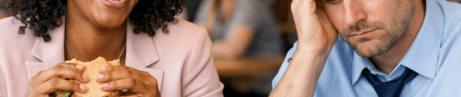 Woman happily eating a sandwich while man looks stressed with burger and laptop